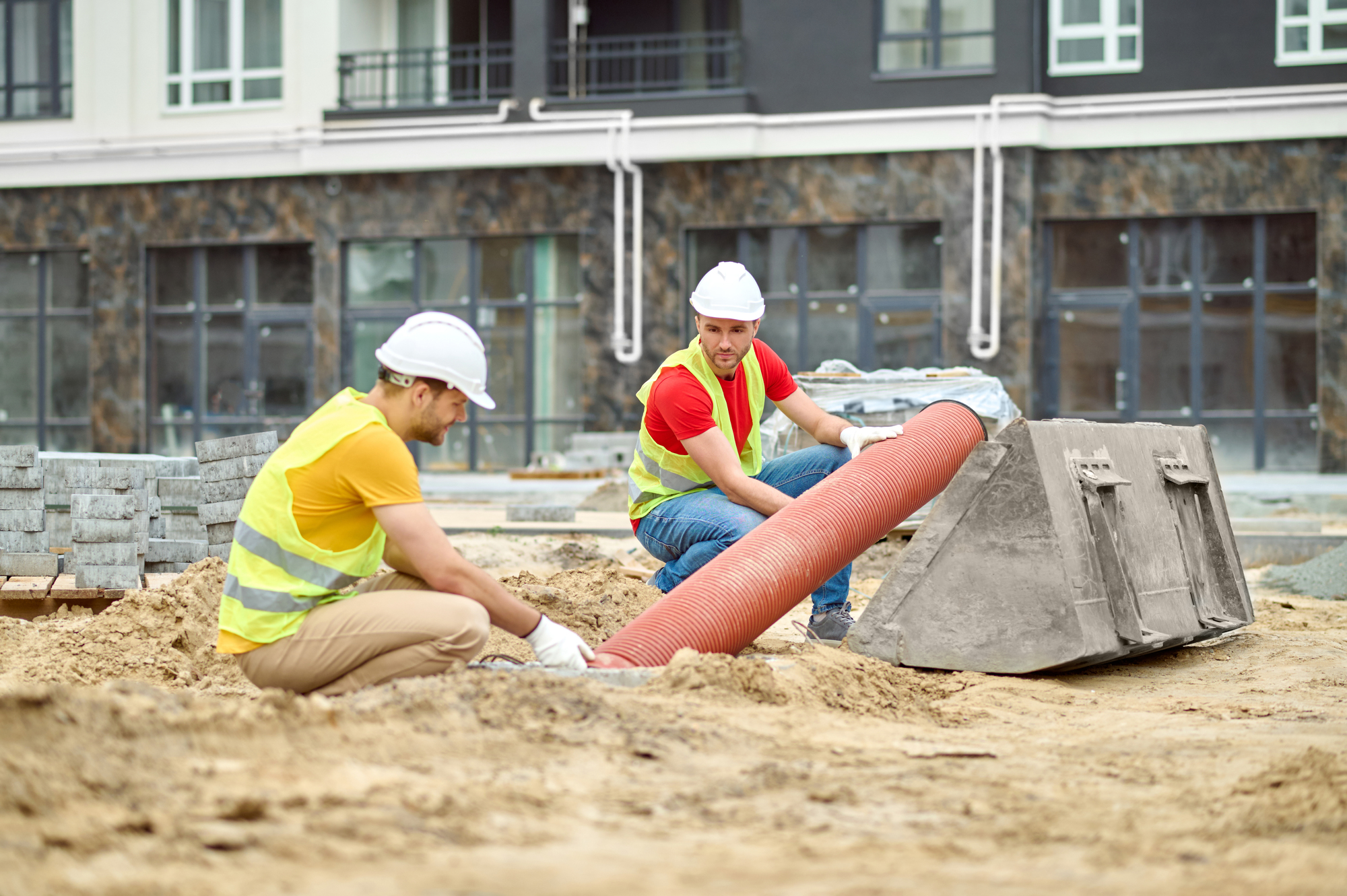 Working mood. Two men in protective helmet and bright vest crouched touching pipe on sand at construction site during day